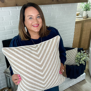 Woman holding a striped pillow in a home setting with a white brick wall and wooden furniture.