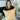 Smiling woman with shoulder-length brown hair holding a yellow floral pillow in front of a white brick fireplace with home decor in the background.