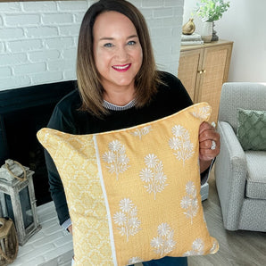 Smiling woman with shoulder-length brown hair holding a yellow floral pillow in front of a white brick fireplace with home decor in the background.